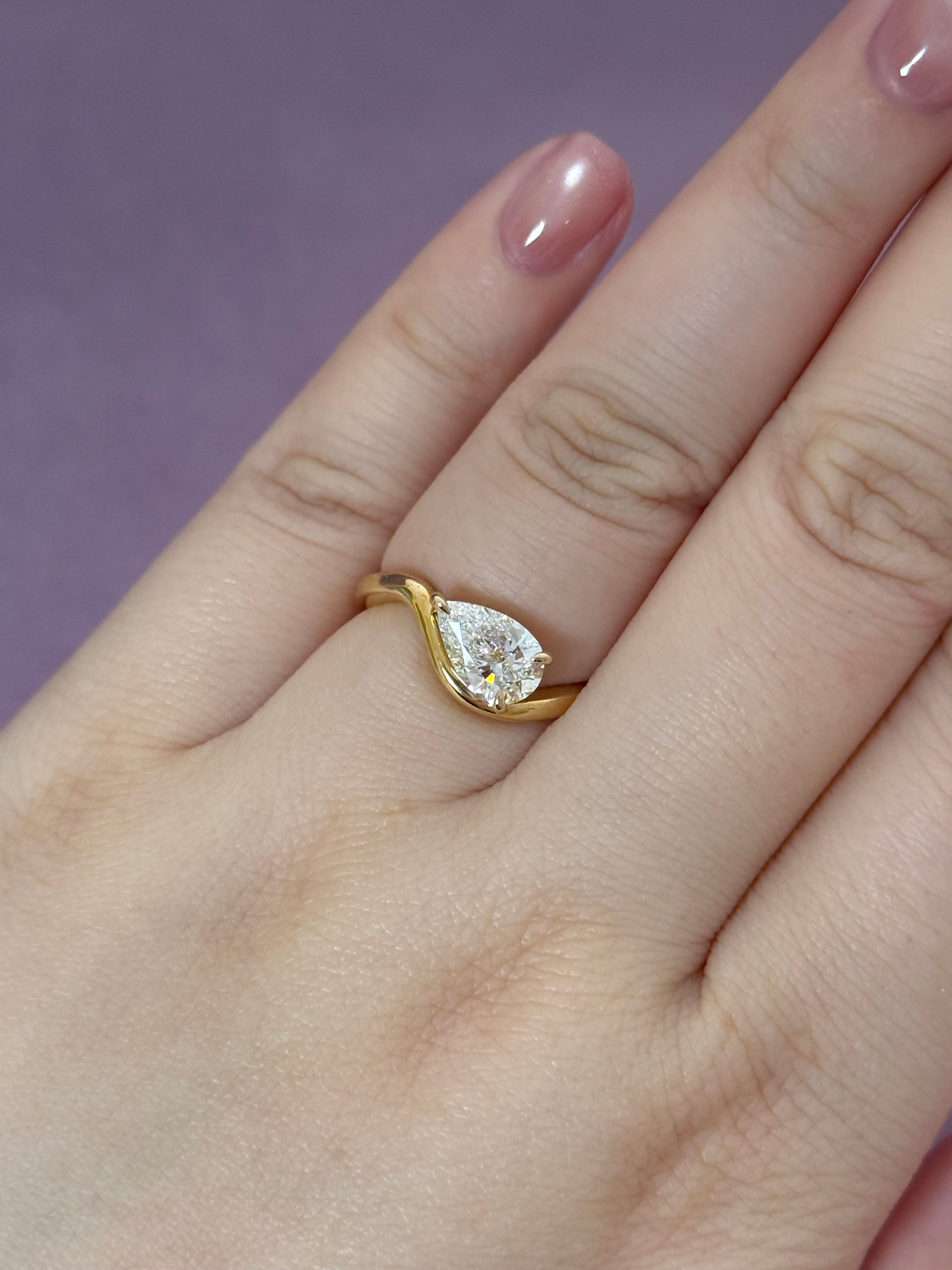 A close-up of a hand wearing a yellow gold ring with a pear-cut clear gemstone, set against a solid purple background.